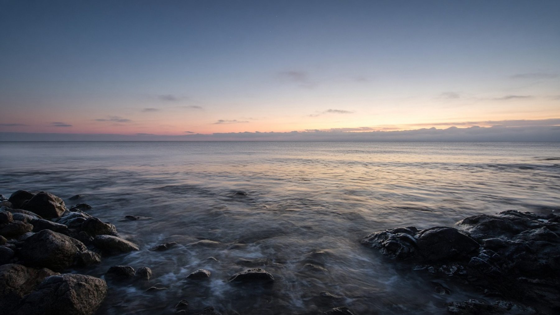 A calm sea at dawn over dark wet stones.